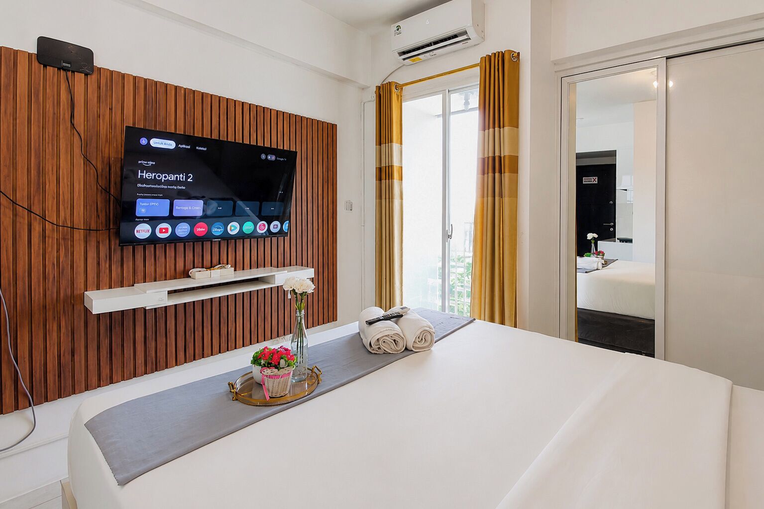 A hotel room with a television mounted on a wooden panel wall, a bedside table with towels and a vase of flowers, and a window with curtains.