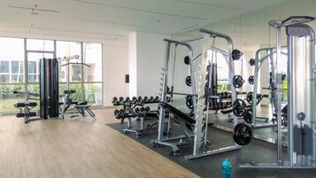 Academia com diversos equipamentos de exercício, janelas amplas e piso de madeira.