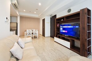 Ein Wohnzimmer mit beiger Couch, einem Flachbildfernseher auf einem Holzschrank, einem Esstisch mit Stühlen und einer Küche im Hintergrund.