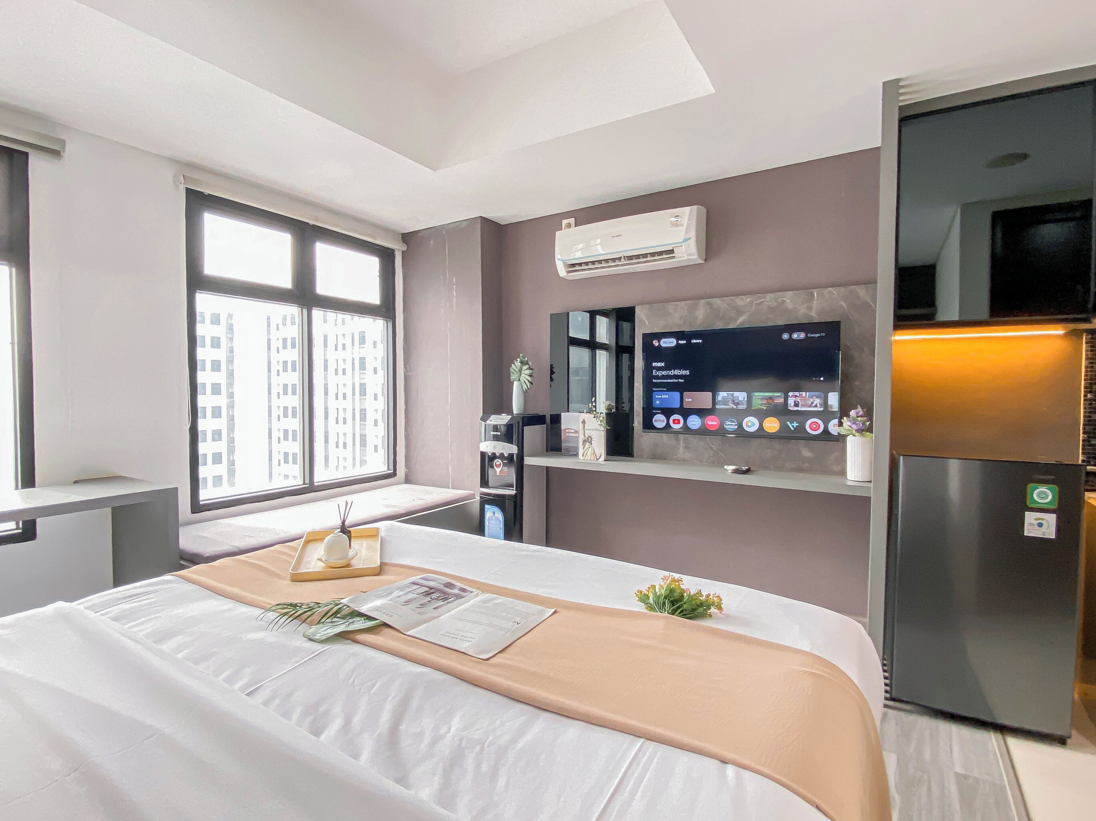 A hotel room with a bed, a TV mounted on the wall, a refrigerator, and a window with a view of buildings.