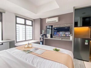 Habitación de hotel con cama, televisor montado en la pared, refrigerador y ventana con vista a edificios.