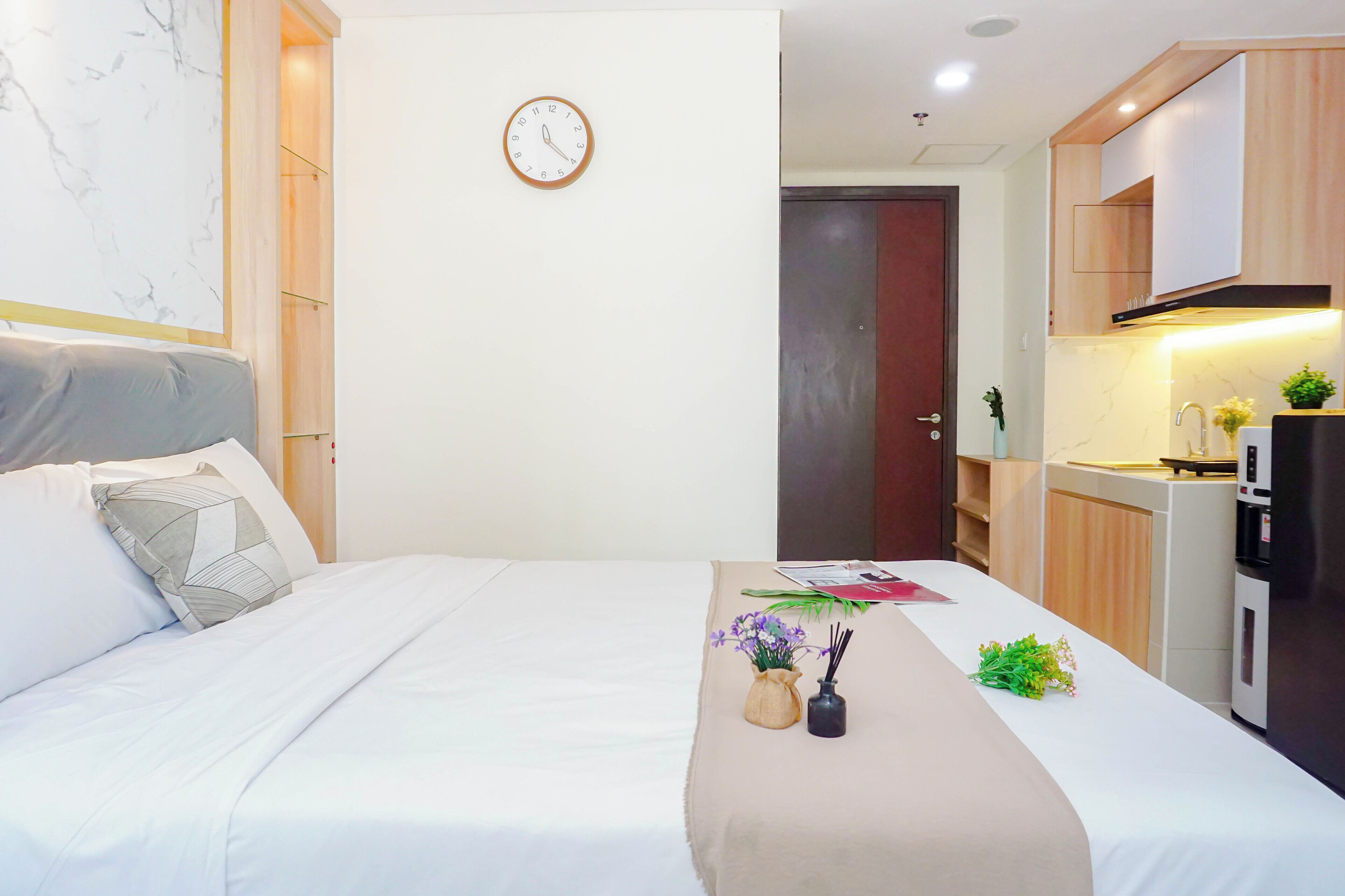 A bedroom with a bed, a bedside table with a vase and flowers, a clock on the wall, and a view into the adjacent kitchen area.