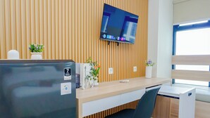 A modern office space with a flat-screen TV mounted on the wall, a wooden desk, a black office chair, and a small refrigerator. - Compact Studio Near President University At Pollux Chadstone Apartment (Cikarang)