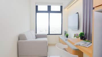 A modern, minimalistic room with a sofa, a desk, a chair, a flat-screen TV, and a large window offering a city view.