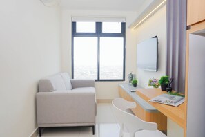 A modern, minimalistic room with a sofa, a desk, a chair, a flat-screen TV, and a large window offering a city view. - 3 Pax Airy 2Br At Pollux Chadstone Apartment (Cikarang)
