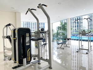 A gym with various exercise machines, a bench, and a view of a swimming pool through large windows. - Modern Haven Studio At Apartment Pollux Chadstone (Cikarang)