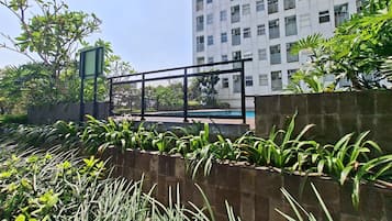 Uma área externa cercada por grades, com piscina, rodeada por vegetação e um edifício alto ao fundo.