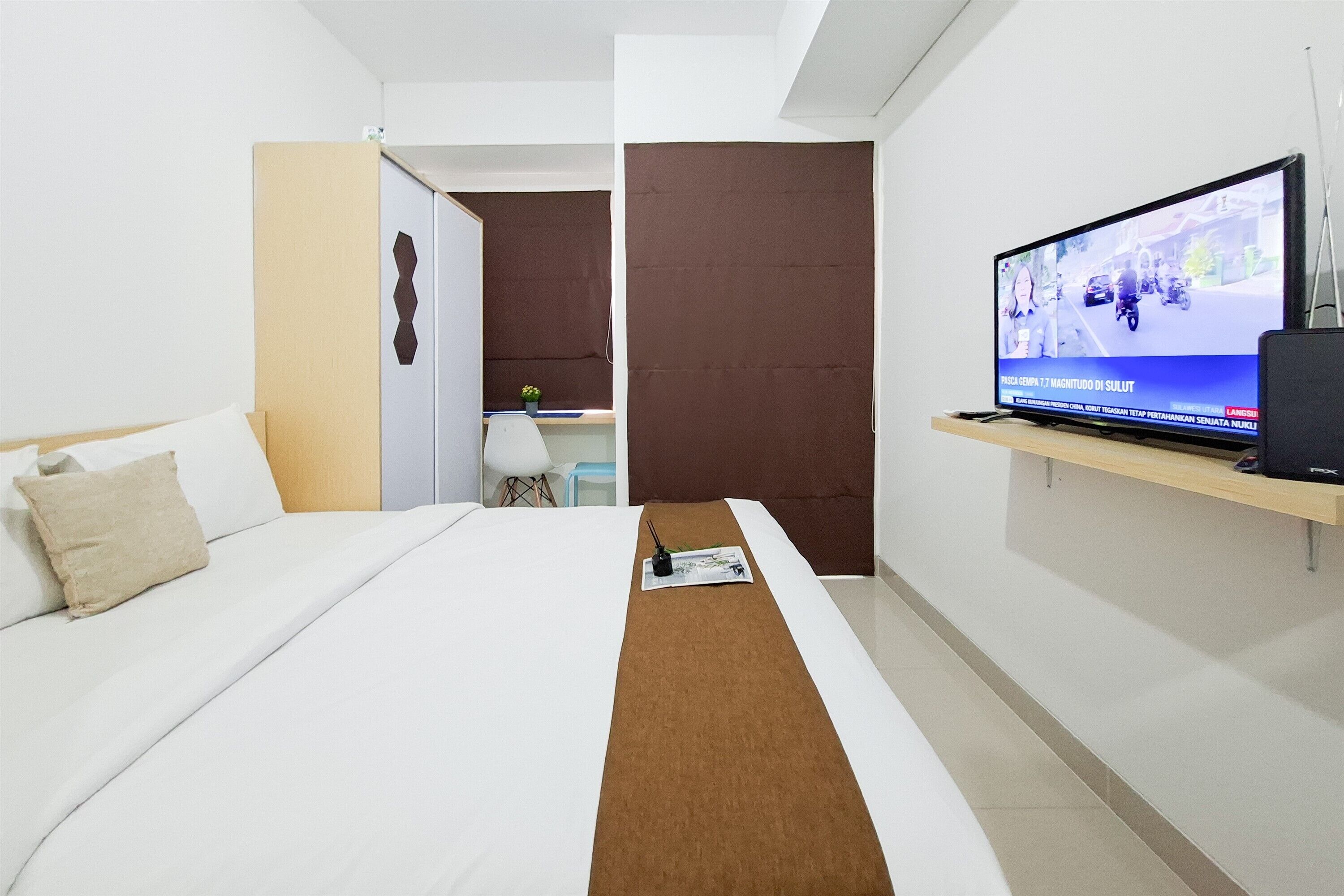 Quarto de hotel com cama, televisão sobre suporte, escrivaninha e uma pequena planta.