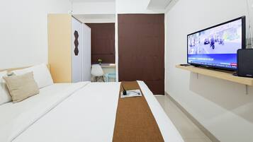 Quarto de hotel com cama, televisão sobre suporte, escrivaninha e uma pequena planta.