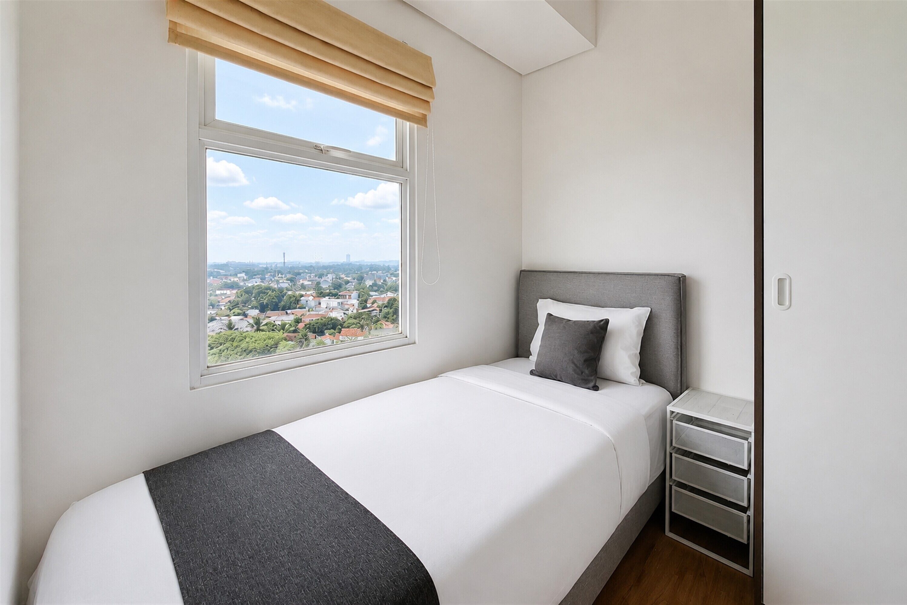 A single bed with a grey headboard and white bedding, a bedside table with drawers, and a window with a view of a cityscape.