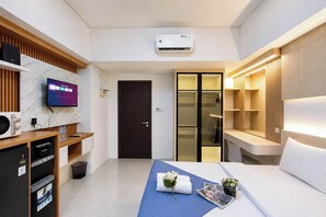 A modern hotel room with a flat-screen TV, air conditioning, a desk with a chair, a bed with a blue bedspread, and a walk-in closet. - Good Deal Studio (No Kitchen) Apartment Skyland City Jatinangor (Sumedang)