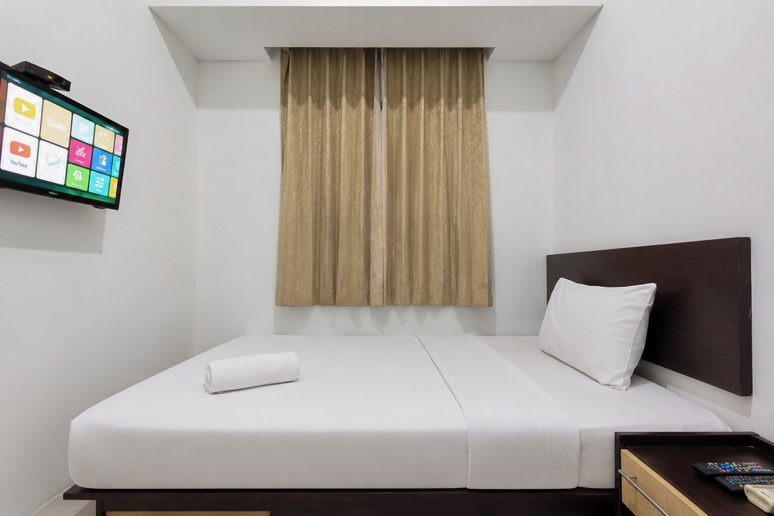 A hotel room with a bed, a TV mounted on the wall, a window with curtains, and a bedside table with a remote control.