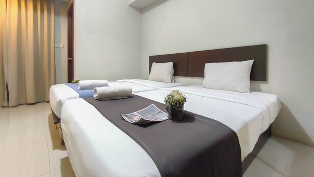 A hotel room with two beds, a large window with curtains, a bedside table with a plant, and a headboard. - Comfy Studio Apartment Skyland City Jatinangor (Sumedang)