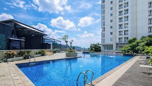 A swimming pool with a clear blue sky and clouds in the background, surrounded by a modern building with balconies and a covered area with plants. - Comfy Studio Apartment Skyland City Jatinangor (Sumedang)