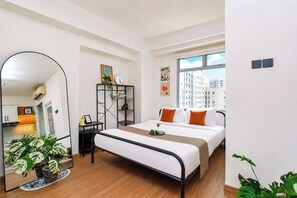 A modern hotel room with a bed, a bedside table, a mirror, a small shelf with plants, and a window with a city view. - Restful And Serene Studio Room At Apartment Green Bay Pluit (Jakarta)