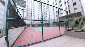 Uma quadra de tênis cercada por uma grade de arame com superfície de jogo vermelha e verde, rodeada por edifícios residenciais altos.