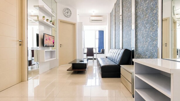 A modern living room with a sofa, a coffee table, and a TV on a stand. There is a clock on the wall and a door leading to another room. - Serene Spacious 2Br At Apartment Benson Supermall Mansion (Surabaya)