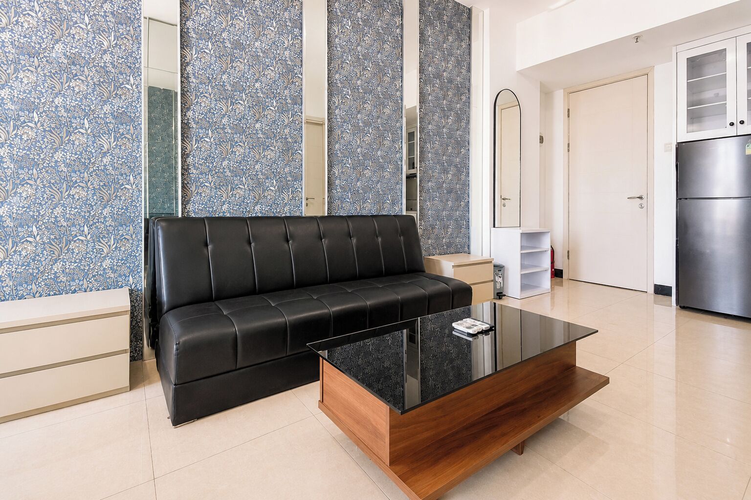 Uma sala de estar moderna com um sofá de couro preto, uma mesa de centro de madeira e uma área de cozinha com geladeira de aço inoxidável.