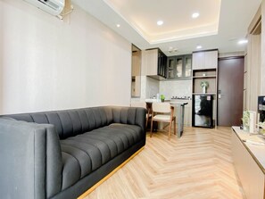A modern living room with a grey sofa, a wooden herringbone floor, and a kitchen area with cabinets and a refrigerator. - Serene Wooden Touch 1Br At Apartemen Daan Mogot City (Jakarta)