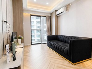 A modern living room with a black sofa, a flat-screen TV, and a large window with a view of a building. - Serene Wooden Touch 1Br At Apartemen Daan Mogot City (Jakarta)