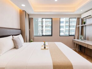 A hotel room with a large bed, a desk, and two windows offering a view of the city. - Serene Wooden Touch 1Br At Apartemen Daan Mogot City (Jakarta)