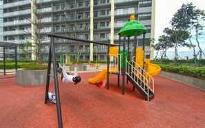 A playground with swings and a slide, surrounded by a red play area and a multi-story building in the background. - Serene Wooden Touch 1Br At Apartemen Daan Mogot City (Jakarta)