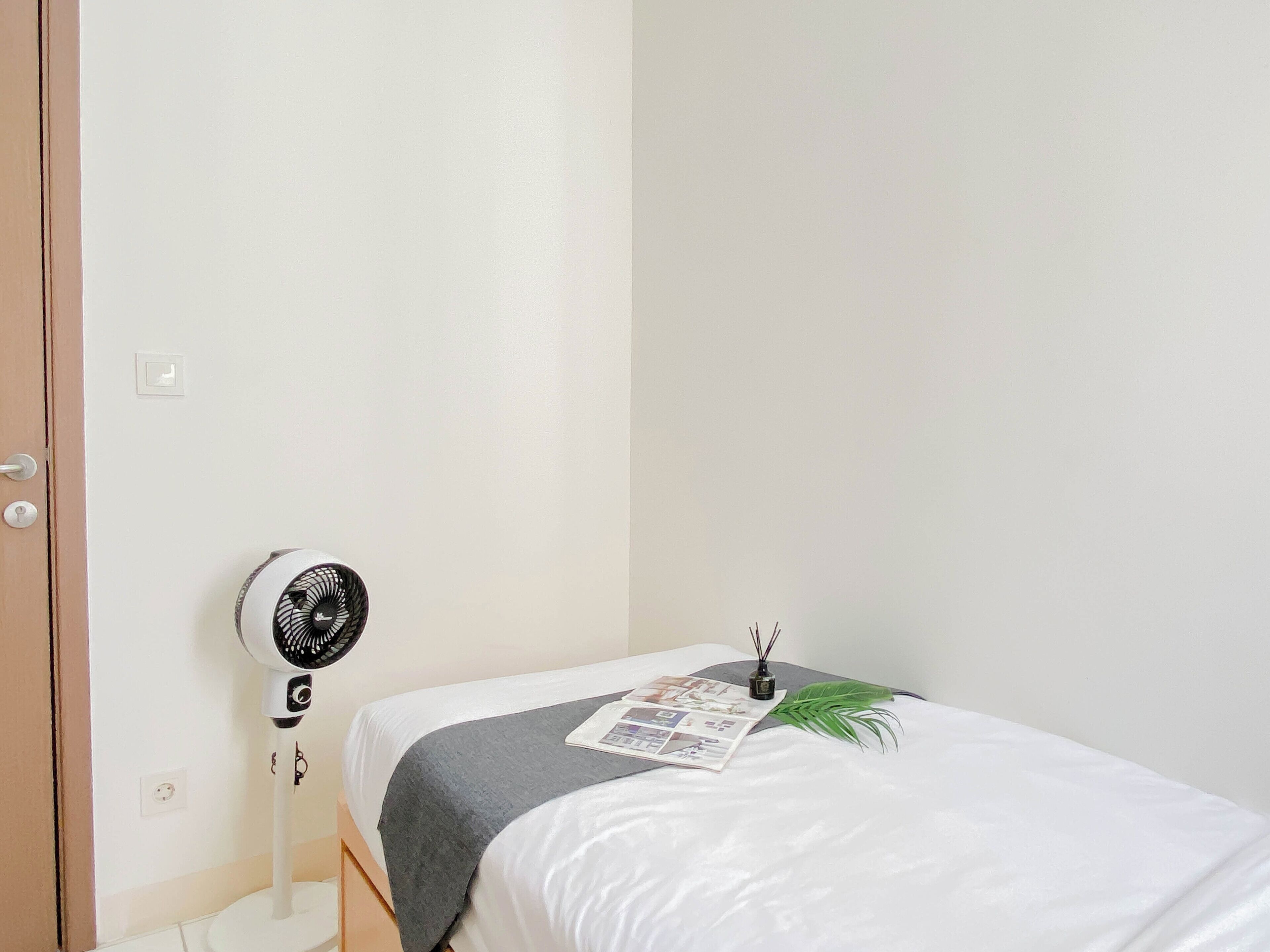 Una cama pequeña y ordenada con sábana blanca y manta gris, un ventilador fijado a la pared y una planta pequeña sobre la cama.