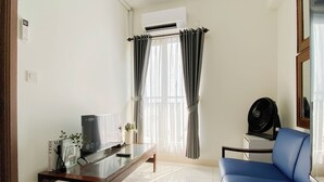 En stue med et sofabord med glasplade, en blå sofa, en sort ventilator og et vindue med gardiner.