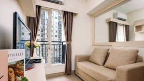 A modern living room with a beige sofa, a flat-screen TV mounted on the wall, a large window with curtains, and an air conditioning unit. - Warm And Homey 2Br Apartment Podomoro Golf View (Depok)