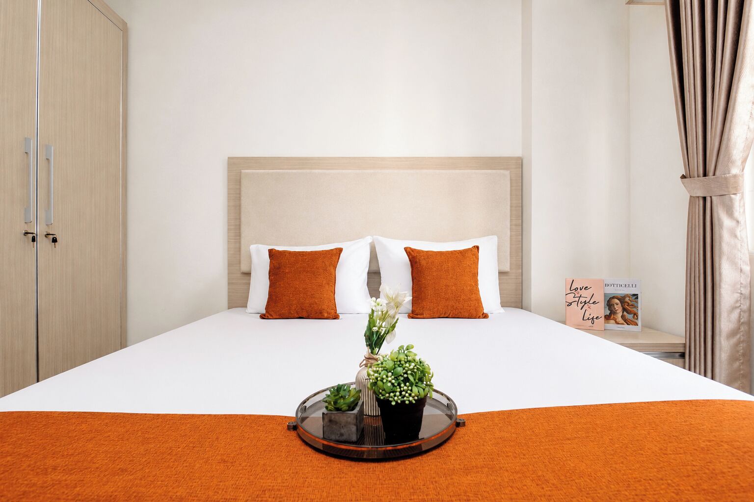 Quarto de hotel com uma cama, duas almofadas, mesa de cabeceira e um livro, e uma janela com cortinas.