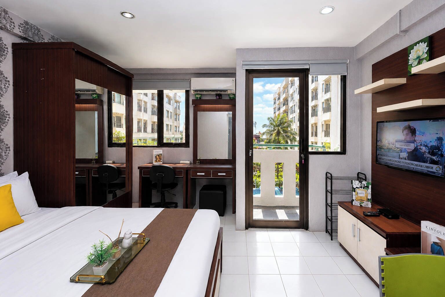 Habitación de hotel con cama, escritorio, silla, televisión y balcón con vistas a edificios.