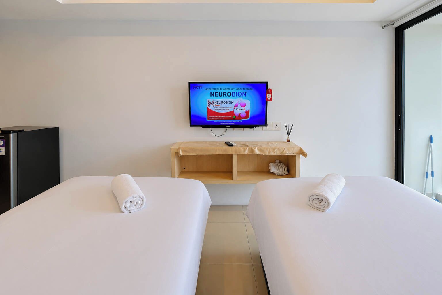 Habitación de hotel con dos camas, un televisor mostrando un anuncio, un minibar y una ventana.