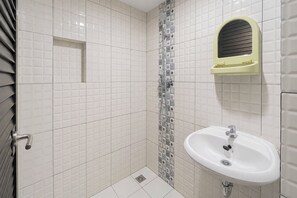 Casa de banho com paredes em azulejo branco, chuveiro com assento amarelo e lavatório branco com torneira cromada.