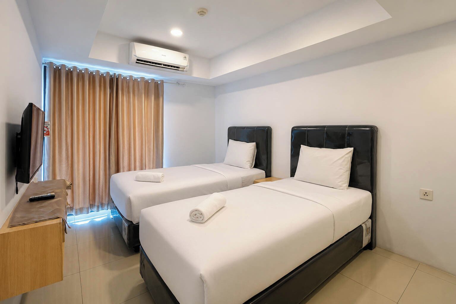 A hotel room with two beds, a TV, an air conditioning unit, and a window with curtains.