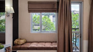 Quarto com janela, um banquinho estofado, luminária e vista para o verde lá fora.