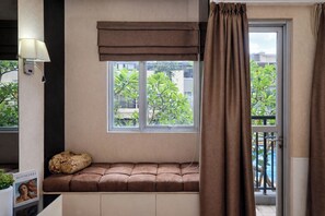 Quarto com janela, um banquinho estofado, luminária e vista para o verde lá fora.