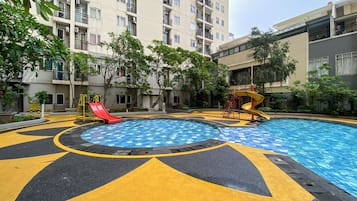 Uma piscina com escorregador e toboágua espiral, cercada por uma área pavimentada com marcações amarelas e pretas, e edifícios residenciais ao fundo.