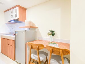 A compact kitchen with a refrigerator, a sink, wooden cabinets, and a small dining table with two chairs. - Homey And Warm Studio Room At Apartment Meikarta District 2 (Cikarang)