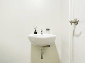 A white bathroom sink with a faucet, a small vase with a plant, and a bottle of reed diffuser. - Bright And Cozy Studio Room At Apartment Meikarta District 2 (Cikarang)