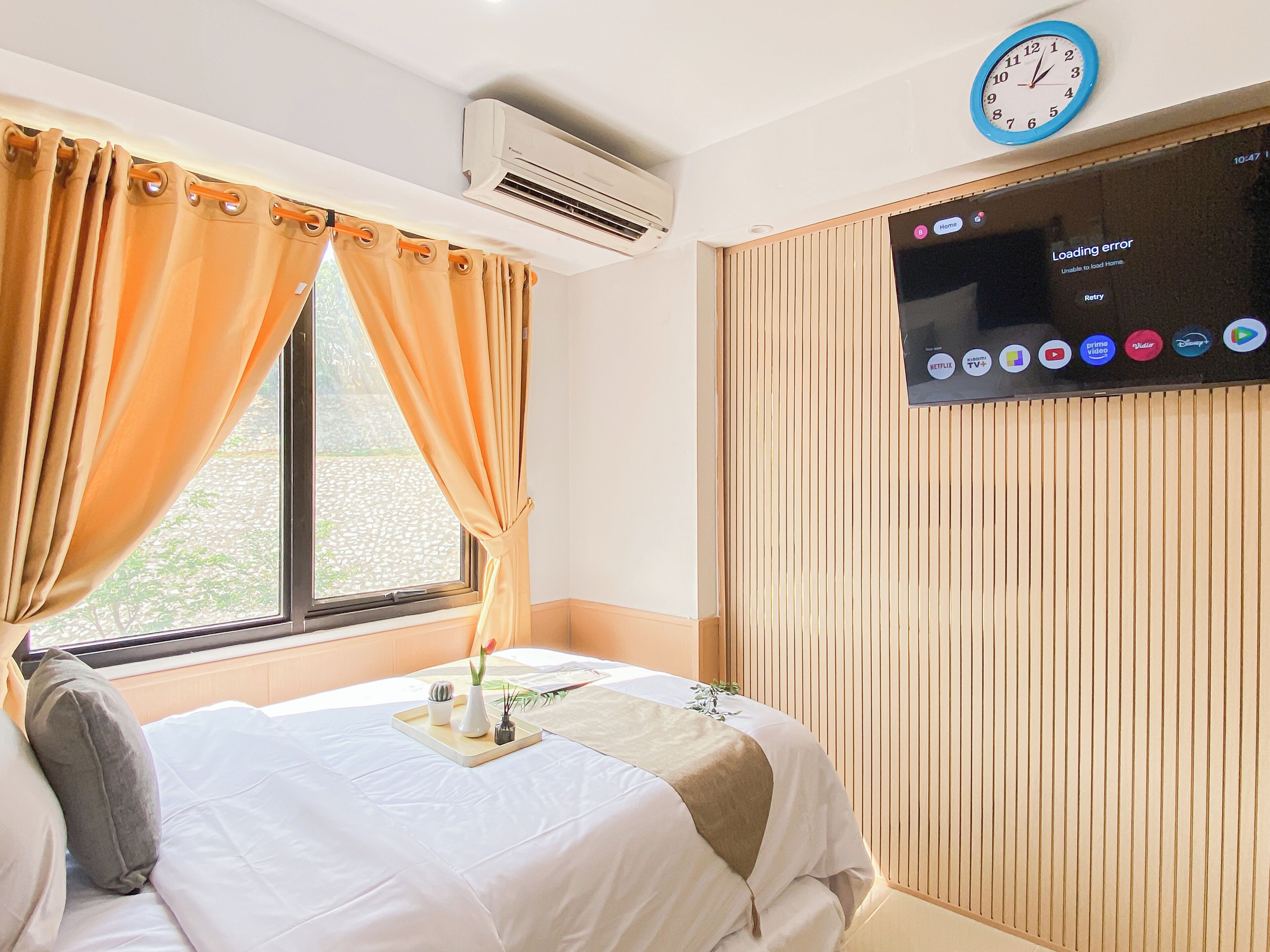 A hotel room with a bed, a window with curtains, an air conditioning unit, a wall-mounted television displaying various channels, and a clock.