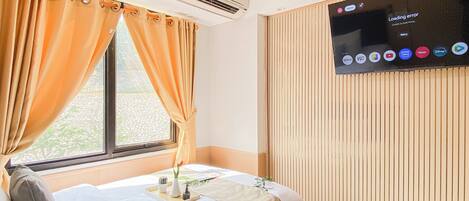 A hotel room with a bed, a window with curtains, an air conditioning unit, a wall-mounted television displaying various channels, and a clock.