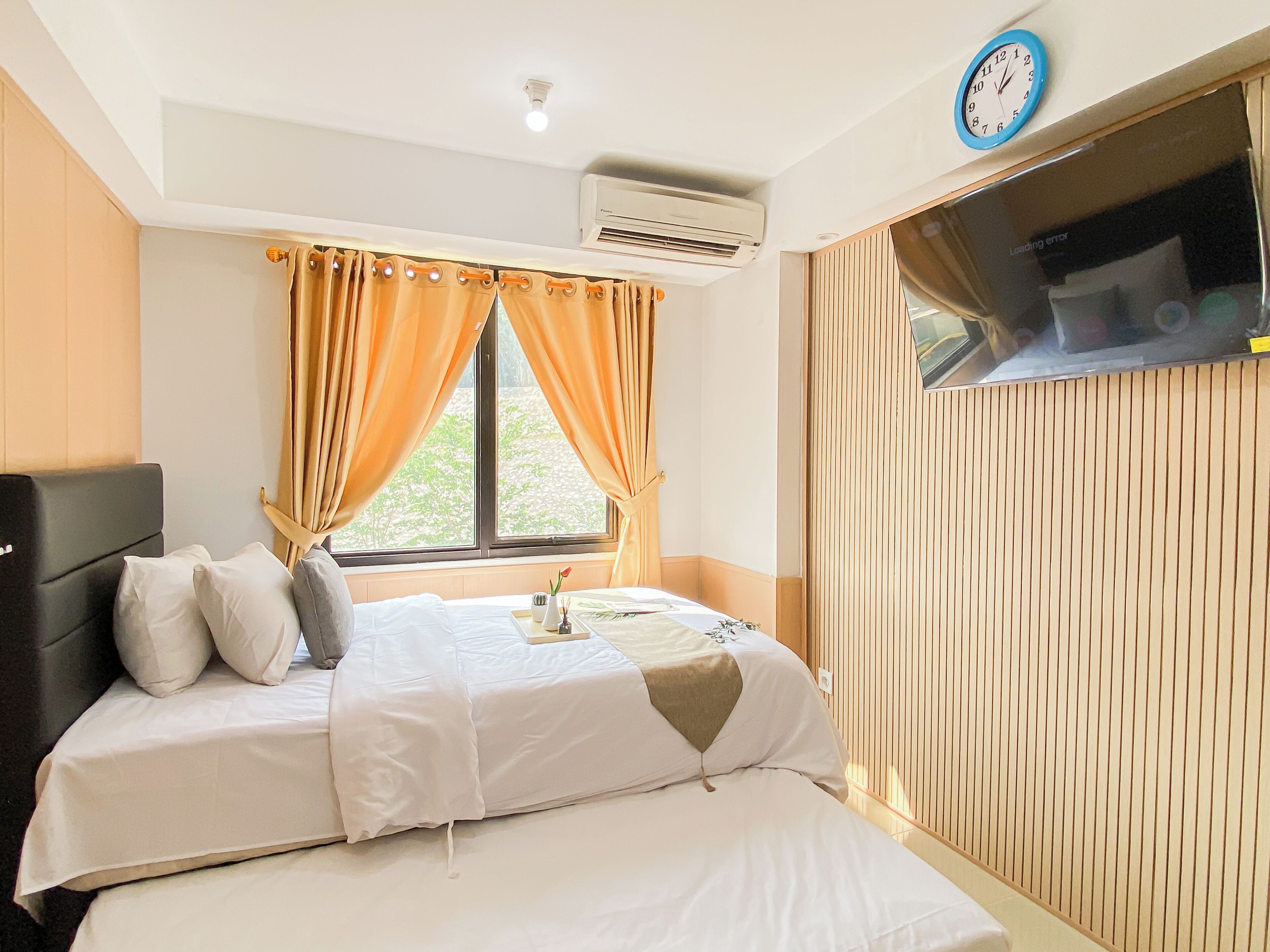 A hotel room with a bed, a bedside table, an air conditioning unit, a clock, and a window with curtains.