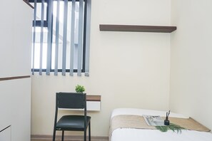 A small, neatly arranged room with a bed, a chair, a window with blinds, and a shelf. - Cozy Stay 2Br At Apartment Meikarta District 2 (Cikarang)