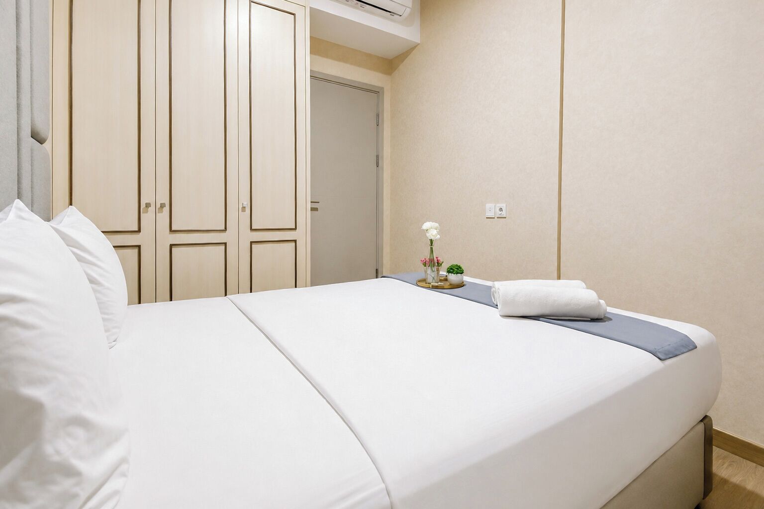 A hotel room with a bed, white curtains, a small table with a vase and a plant, and a closed door.
