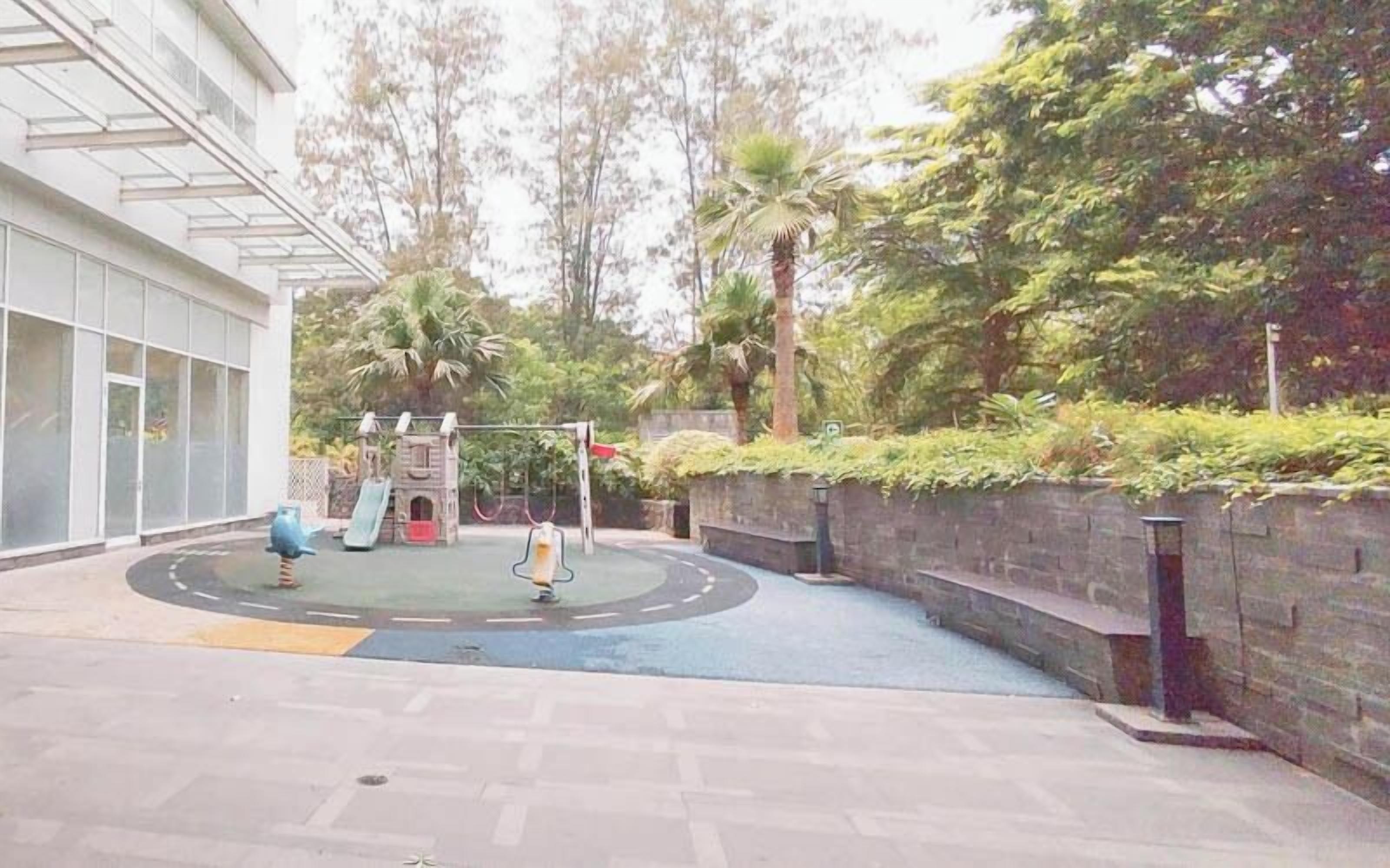 Un parque infantil con tobogán y columpios, rodeado por un edificio con ventanas amplias y una zona pavimentada.