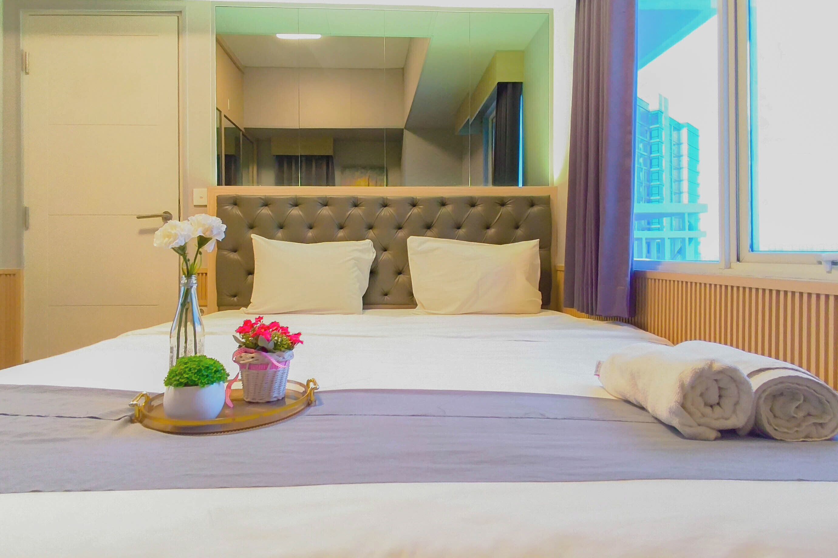 A hotel room with a large bed, a headboard, a window with a view of buildings, and a tray with flowers and a plant.