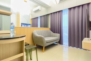 Ein Hotelzimmer mit einer Couch, einem kleinen Tisch, einer Wasserflasche, einem Fernseher und einem Fenster mit Vorhängen.