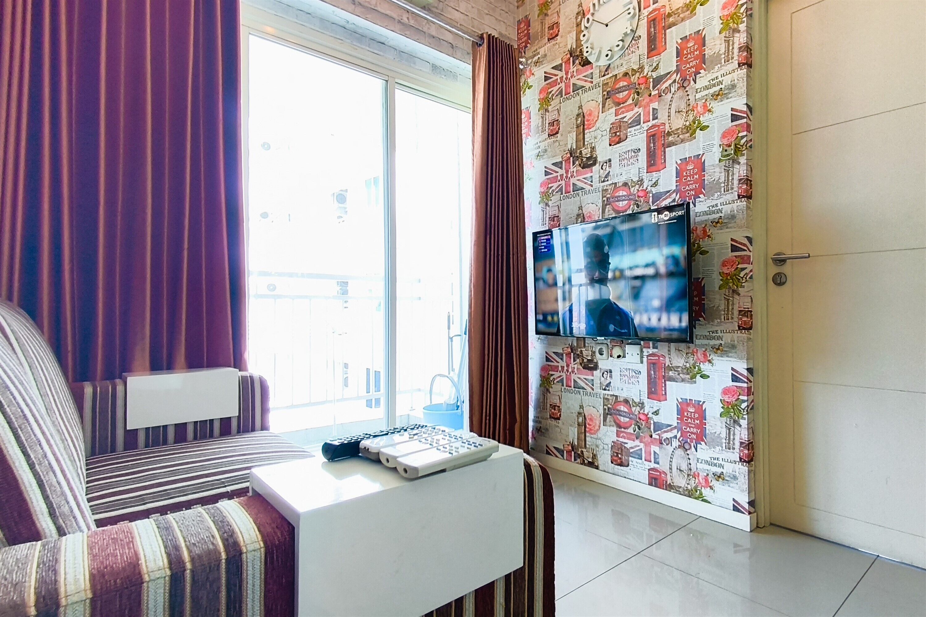 Habitación de hotel con una cama, un escritorio con un teclado, un televisor montado en la pared y una ventana con cortinas.