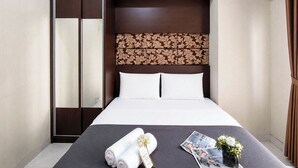 A hotel room with a bed, a wardrobe, a headboard with a floral pattern, and a small table with a magazine and a flower arrangement. - Serene 2Br Apartment Newton Residence Bandung (Bandung)