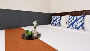 Hotellihuone, jossa on sänky, yöpöytä maljakolla ja kasveilla sekä sängynpääty, jossa on kuvioituja koristetyynyjä.